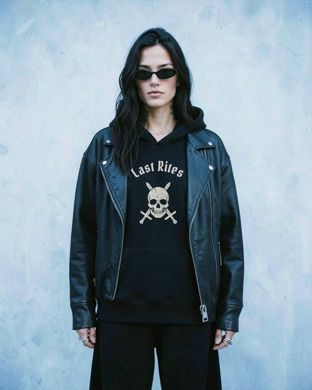 A woman with long dark hair, small black oval sunglasses and neck tattoos, in the black Crest Hoodie under an open leather moto jacket, against a pale icy blue wall.