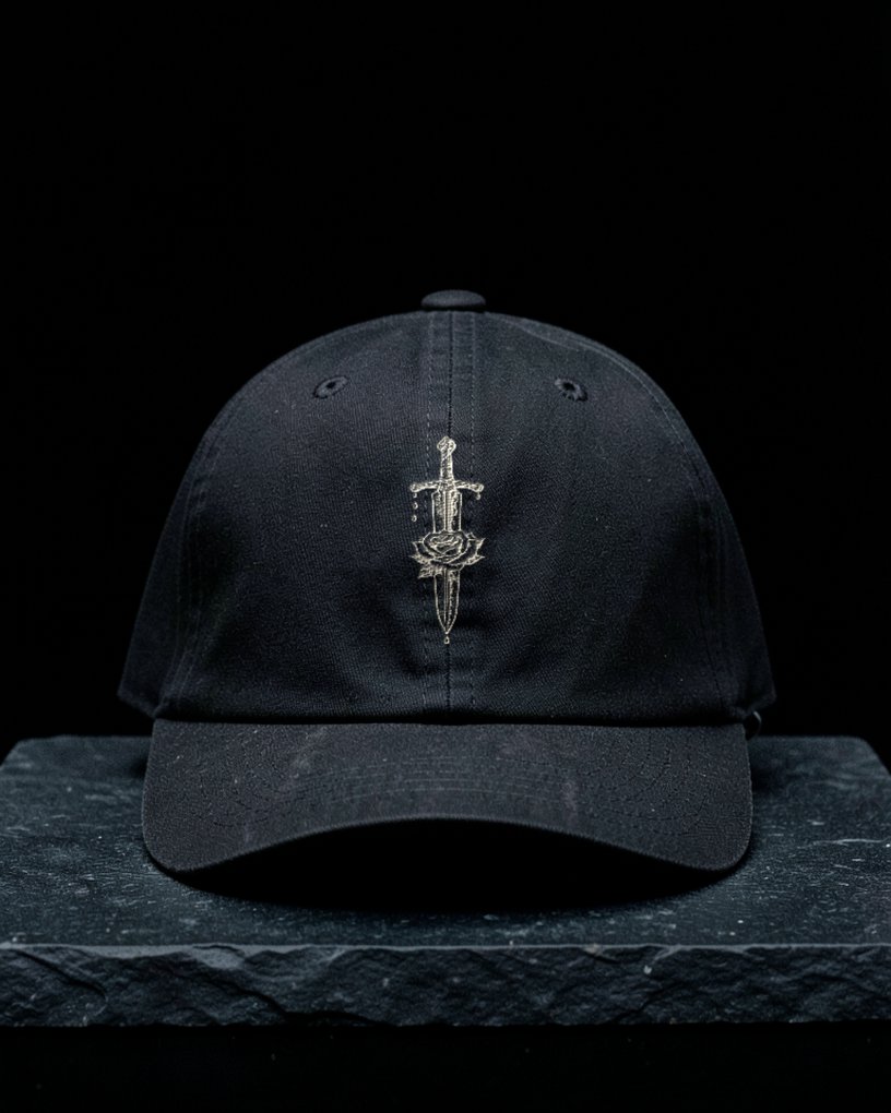 A washed black low-profile six panel cap embroidered in bone thread with a tattoo-flash dagger piercing a rose.