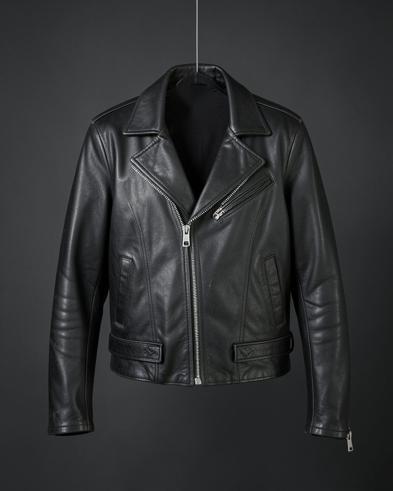 A black leather motorcycle jacket with an asymmetric silver zip and belted waist, hanging on a thread against charcoal.